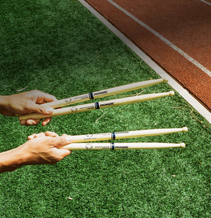 Two hands holding drumsticks on a grassy surface with a track in the background