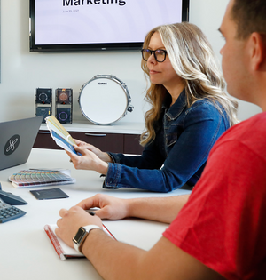 Two people working at a desk with a laptop and color swatches, with a television screen displaying 'Marketing' in the background.